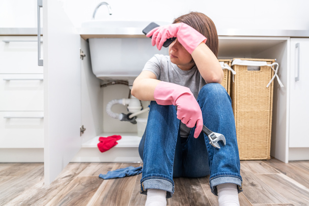 Woman sitting near leaking sink in laundry room holding adjustable wrench - 写真・画像