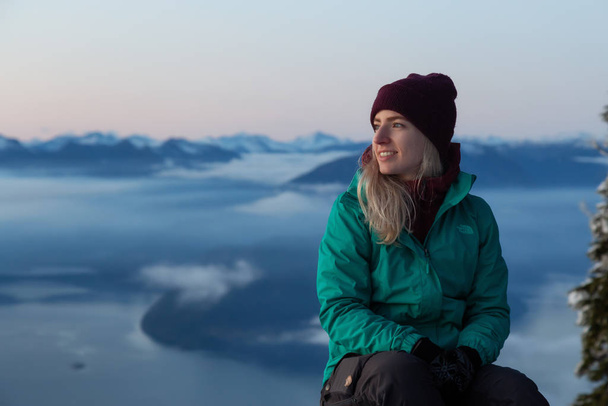 Vancouver, BC, Canada - November 25, 2019: Adventurous Blond Caucasian Girl sitting on top of a snow covered mountain during a colorful winter sunset. - Foto, immagini