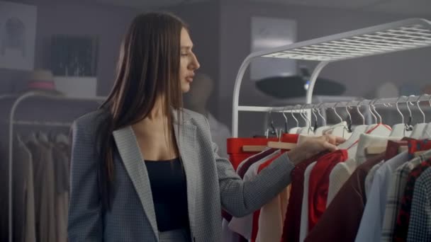 junge schöne Mädchen in einem Bekleidungsgeschäft suchen Kleider und Pullover auf der Theke. vom Kleiderbügel zu nehmen und sich lächelnd zu stellen - Filmmaterial, Video