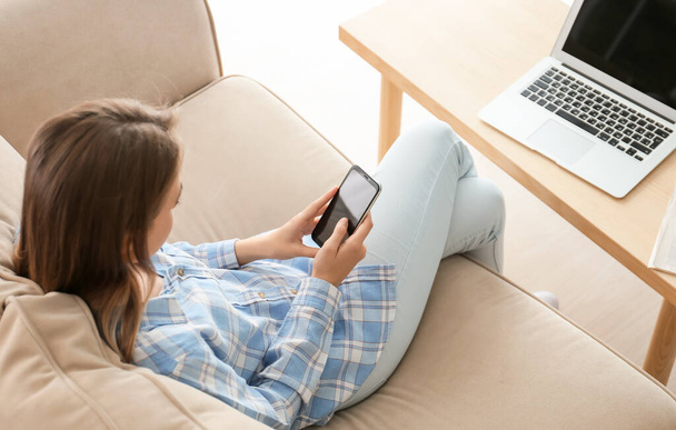 Nuori nainen, jolla on matkapuhelin istuu sohvalla kotona
 - Valokuva, kuva