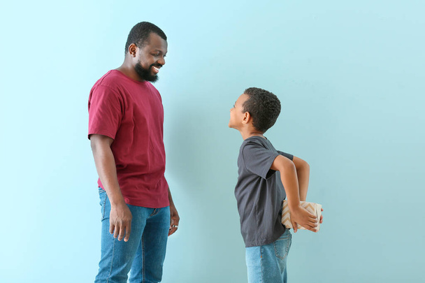 Little African-American boy hiding gift from his father behind back against color background - Photo, Image