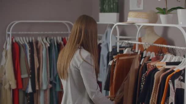Young beautiful girl in a clothing store looking dresses and sweaters on the counter. To take from a hanger and to put to itself smiling - Filmagem, Vídeo