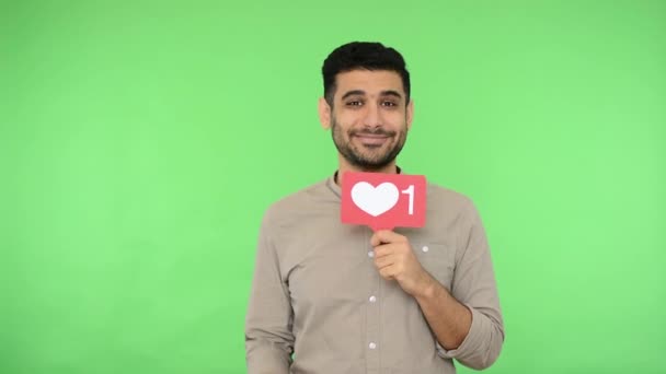 Cheerful handsome man with bristle in shirt winking playfully, pointing to you and showing social media heart Like icon, subscribe and follow, love content. studio shot, green background, chroma key - Filmagem, Vídeo