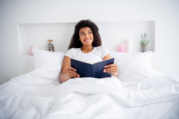 Photo of pretty dark skin lady lying sheets bed white blanket hold book reading novel spend free time dreaming about main character weekend morning wear pajama room indoors - Fotoğraf, Görsel