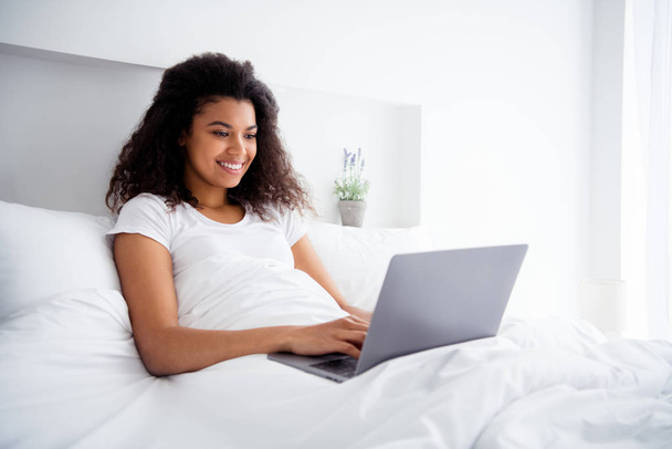 Photo of charming curly dark skin lady lying linen bed covered white blanket hold notebook writing corporate report working weekend saturday morning wear pajama room indoors - Photo, Image