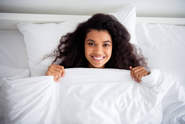 High angle above view photo of pretty dark skin lady lying sheets bed covered white blanket enjoy weekend saturday morning toothy smiling wear pajama room indoors - Fotografie, Obrázek