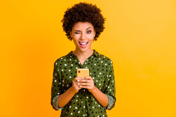 Portrait of excited crazy afro american girl hold smart phone use read social network news wear stylish green bright clothing isolated over yellow color background - Photo, image