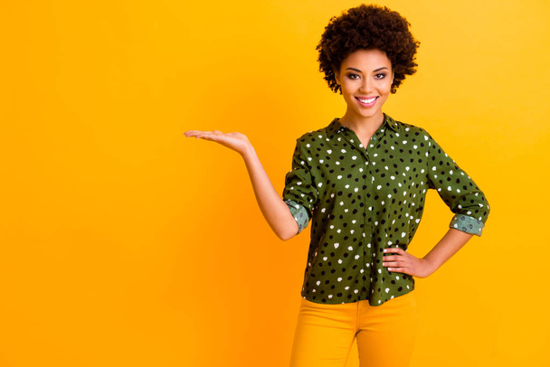 Photo of beautiful dark skin wavy lady holding novelty product on open palm empty space sales manager wear green dotted shirt blouse pants isolated yellow color background - Fotografie, Obrázek