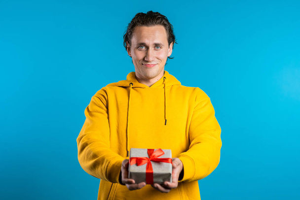 Handsome man holding christmas gift and gives it by hands it to camera. He is happy, smiling. Guy on blue background. Positive holiday shot - Photo, image