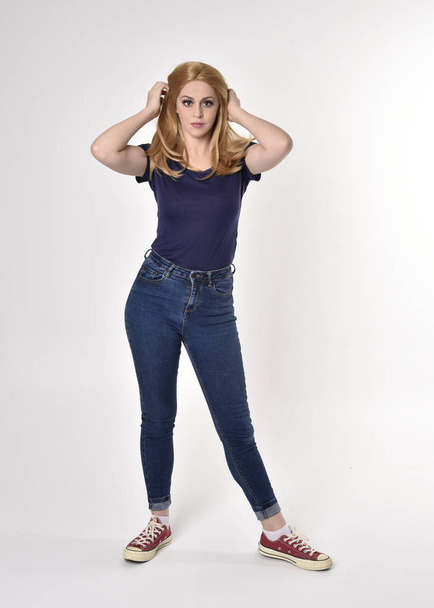 Full length portrait of a pretty blonde girl wearing casual blue shirt, denim jeans and sneakers. Standing pose on a studio background. - Фото, изображение