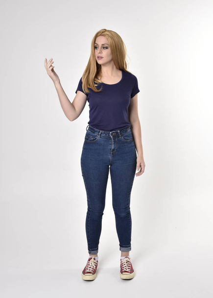 Volledig portret van een mooi blond meisje met casual blauw shirt, spijkerbroek en sneakers. Staande houding op een studio achtergrond. - Foto, afbeelding