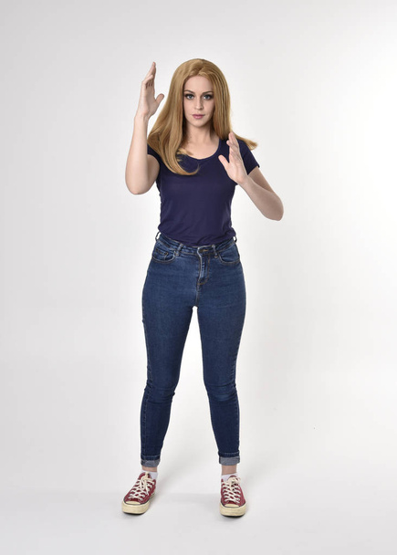 Dlouhý portrét hezké blondýnky v neformální modré košili, džínách a teniskách. Stojící póza na pozadí studia. - Fotografie, Obrázek