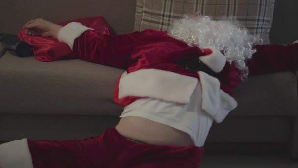 Drunk old man with false white beard in the costume of Santa Claus falls out of bed at home and confused looking around. Bad Santa. Alcoholism, depression, negativism, loneliness - Materiał filmowy, wideo