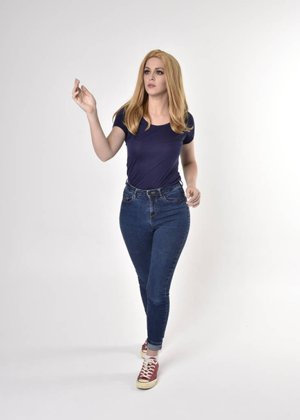 Full length portrait of a pretty blonde girl wearing casual blue shirt, denim jeans and sneakers. Standing pose on a studio background. - Zdjęcie, obraz