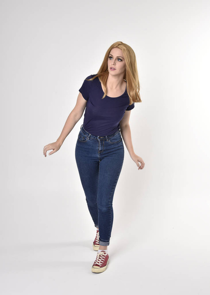 Full length portrait of a pretty blonde girl wearing casual blue shirt, denim jeans and sneakers. Standing pose on a studio background. - 写真・画像