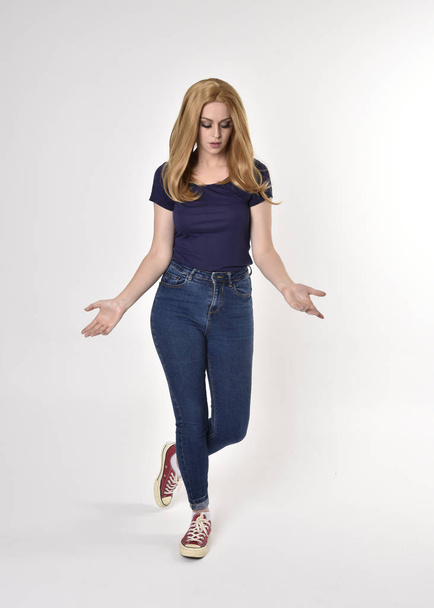 Full length portrait of a pretty blonde girl wearing casual blue shirt, denim jeans and sneakers. Standing pose on a studio background. - Fotó, kép