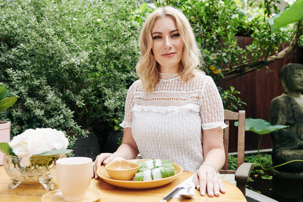 Portrait d'une jolie jeune femme blonde souriante assise à table dans un café extérieur et mangeant le déjeuner
 - Photo, image