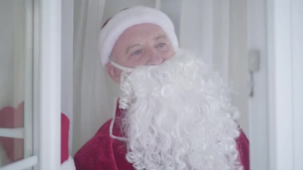 Portrét staříka v kostýmu Santa Clause otvírajícího okno, volajícího svému příteli a naslouchajícího odpovědi, pak mávajícího rukou. Koncept šťastných svátků, tradice, Vánoce. - Záběry, video
