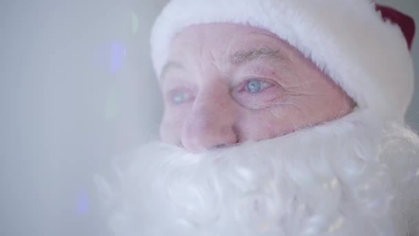 Close-up portret van de oude man met valse baard in kostuum van de kerstman huilend voor nieuwjaarsboom. Tranen verschijnen op de mensenogen. Concept van verdriet, verdriet, gebrek aan feestelijke stemming - Video