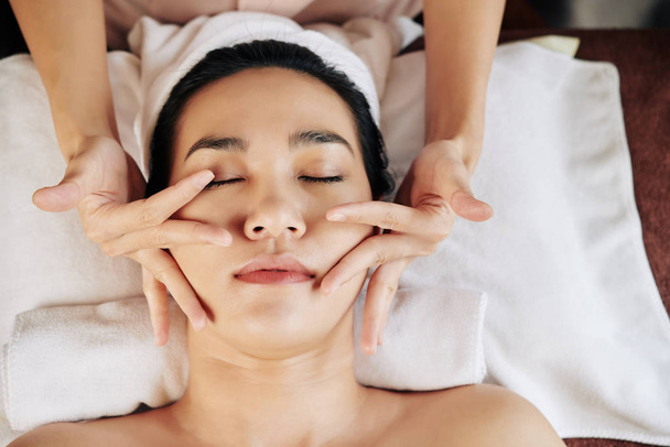 Giovane donna asiatica sdraiata sul tavolo da massaggio e godendo di massaggio facciale in clinica di bellezza, vista dall'alto
 - Foto, immagini