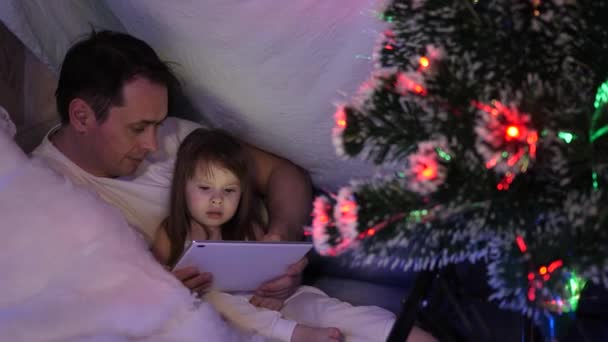 Father and daughter on Christmas evening, play and watch cartoons on tablet, in a childrens room in a tent with garlands. baby and dad are playing in room. concept of happy childhood and family. - Metraje, vídeo