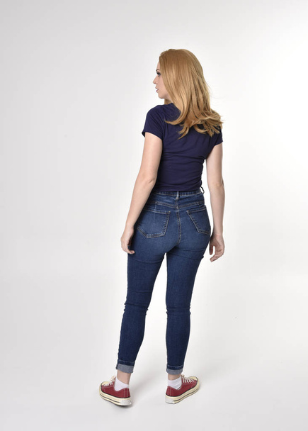 Full length portrait of a pretty blonde girl wearing casual blue shirt, denim jeans and sneakers. Standing pose, walking away from the camera, on a studio background. - Fotó, kép