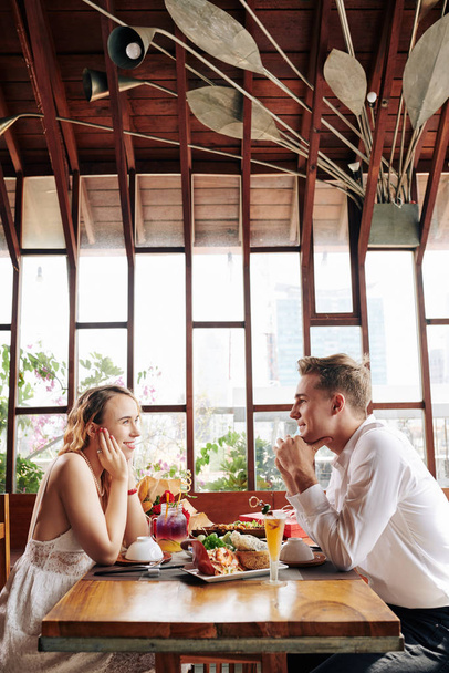 Happy young couple in love looking at each other and smiling when enjoying romantic date in cafe - 写真・画像