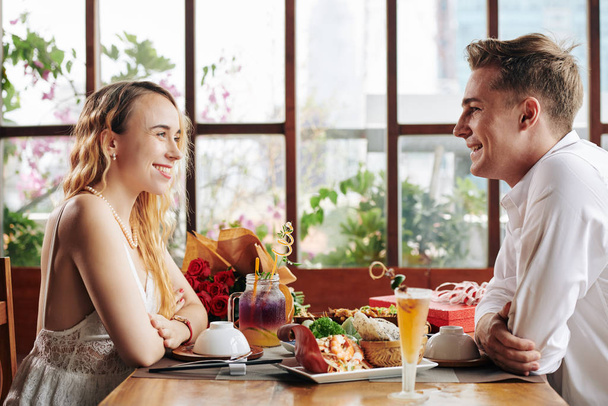 Χαρούμενο νεαρό καυκάσιο ζευγάρι που περνάει το πρώτο του ραντεβού στο καφέ και απολαμβάνει νόστιμο φαγητό και συζήτηση - Φωτογραφία, εικόνα