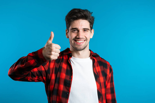 Positive young man smiles to camera. Hipster guy showing thumb up sign over blue background. Winner. Success. Body language. - 写真・画像