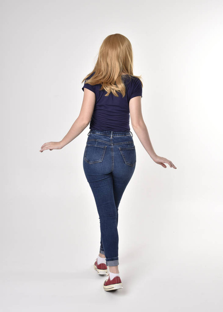 Dlouhý portrét hezké blondýnky v neformální modré košili, džínách a teniskách. Stojící póza, odchází od kamery, na pozadí studia. - Fotografie, Obrázek