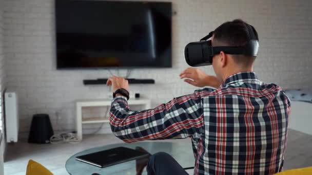 Young modern caucasian man in a VR-headset. Back view. Sits on a sofa in a cozy smart apartment near the window. Plays a computer game in augmented reality. Smart House. Plaid shirt. Camera span - Footage, Video