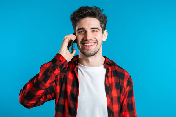 Young man in bright colorful wear talking on mobile cell phone on blue background. Trendy guy have conversation. Smartphone, technology concept. - Photo, image
