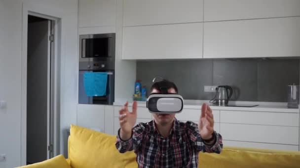 Nuori valkoihoinen mies VR-kuulokkeissa. Etunäkymä. Istuu sohvalla viihtyisässä älykkäässä huoneistossa keittiön taustalla. Pelaa tietokonepeliä täydennetyssä todellisuudessa. Smart Housessa. Plaid-paita
. - Materiaali, video