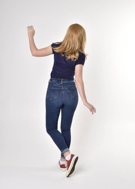 Portrait complet d'une jolie fille blonde portant une chemise bleue décontractée, un jean en denim et des baskets. pose debout, s'éloignant de la caméra, sur un fond de studio
. - Photo, image