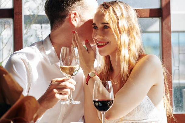 Jongeman fluistert liefdeswoorden tegen zijn lachende vriendin als ze wijn drinken in restaurant - Foto, afbeelding