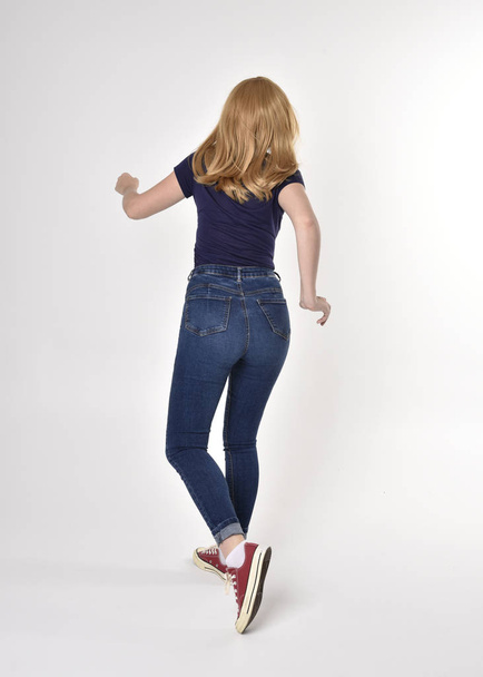 Full length portrait of a pretty blonde girl wearing casual blue shirt, denim jeans and sneakers. Standing pose, walking away from the camera, on a studio background. - Photo, image