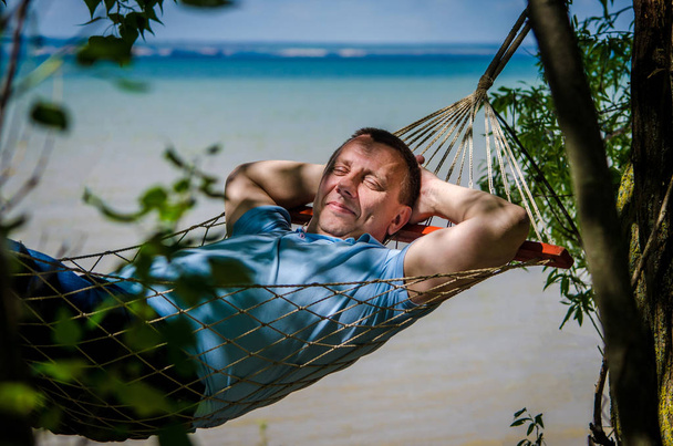 A man in a blue t-shirt swings in a hammock on beach, smiles in shade of trees. Vacation on a tropical island. The concept of relaxation, pleasure, pleasant bliss while traveling. - Foto, Imagem