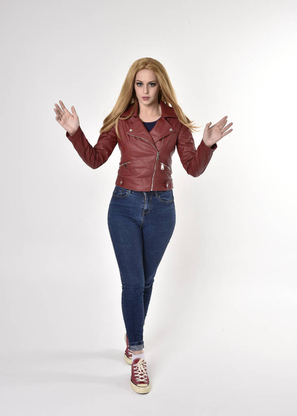 Dlouhý portrét hezké blondýnky v červeném koženém saku džíny a tenisky. Stojící póza na pozadí studia. - Fotografie, Obrázek