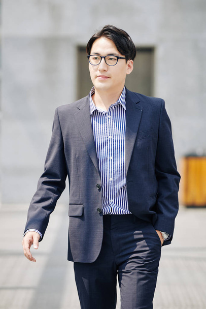Уверенный азиатский бизнесмен в костюме и очках, гуляющий по улице
 - Фото, изображение