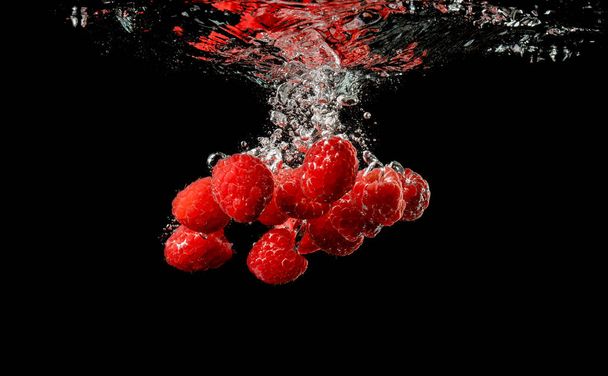 Frambuesas rojas caen bajo el agua con un chapoteo, aisladas sobre fondo negro
. - Foto, Imagen
