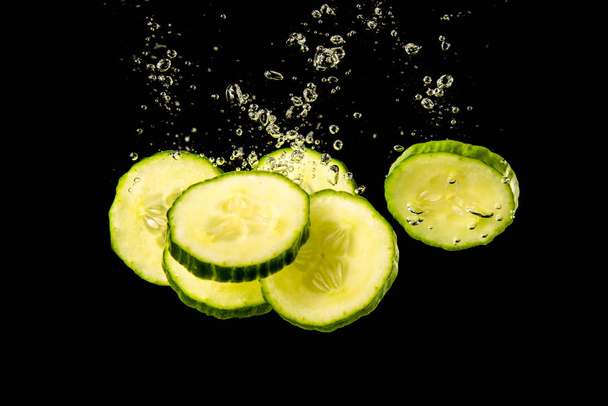 Gesneden komkommer spetterend water geïsoleerd op zwarte achtergrond. Gezondheid en cosmetica. - Foto, afbeelding