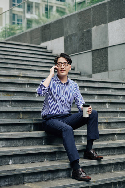 Beau jeune homme asiatique dans des lunettes assis sur les marches, boire du café à emporter et appeler au téléphone
 - Photo, image