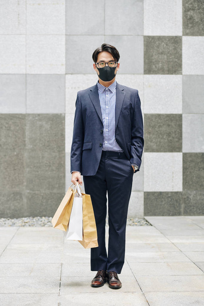 Täyspitkä muotokuva Aasian yrittäjä kasvot naamio seisoo rautatieasemalla ostoskassit
 - Valokuva, kuva