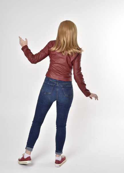 Ganzkörperporträt eines hübschen blonden Mädchens in roter Lederjacke, Jeans und Turnschuhen. im Stehen, mit dem Gesicht zur Kamera, auf Studiohintergrund. - Foto, Bild