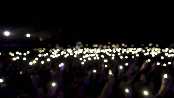 Defocused spectators in hall with lights waving in the dark. Action. Audience holding phones with shining flashlights during the concert, romantic atmosphere. - Photo, Image