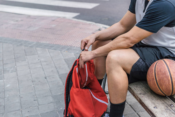 Ένας αθλητής ετοιμάζει κάτι στο κόκκινο σακίδιό του ενώ κάθεται σε ένα παγκάκι - Φωτογραφία, εικόνα