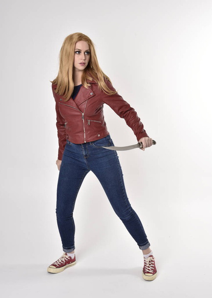 Volledig portret van een mooi blond meisje in een rood leren jasje met spijkerbroek en sneakers. Staande houding, met een dolk, op een studio-achtergrond. - Foto, afbeelding