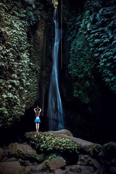 Mujer parada en la roca, practicando yoga. Mujer joven levantando brazos con namaste mudra cerca de la cascada. Cascada Leke Leke, Bali. Vista desde atrás
. - Foto, imagen
