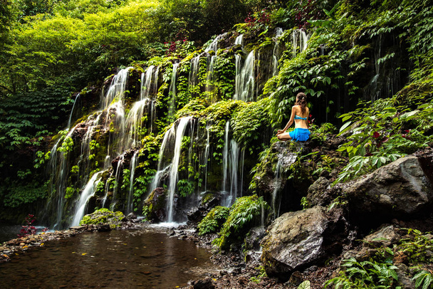 Jonge vrouw zittend op de rots, yoga beoefenend bij de waterval. Handen in de gynaecoloog. Banyu Wana Amertha waterval, Bali. Zicht van achteren. - Foto, afbeelding
