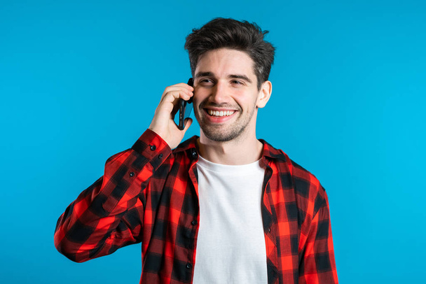 Young man in bright colorful wear talking on mobile cell phone on blue background. Trendy guy have conversation. Smartphone, technology concept. - Foto, afbeelding